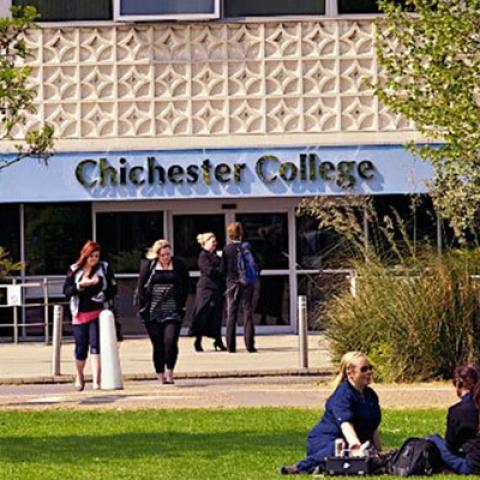 A view of the outside of the main entrance to Chichester College with students walking and sitting on the grass