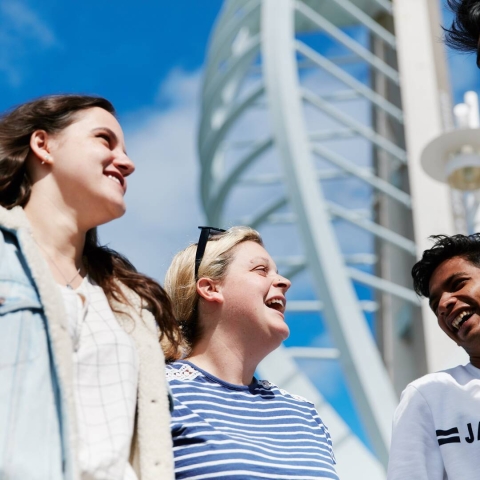 A group of people laughing together, stood against a blue sky and the Spinnaker Tower. Overlay text reads: Research Futures Human Connections and Flourishing