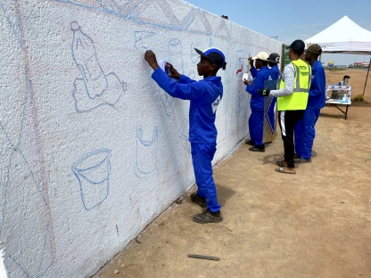 A group of people in South Africa in the early stages of drawing a wall mural of recyclable items. The group are drawing the items before painting them. You can see a bottle and a bucket have already been drawn