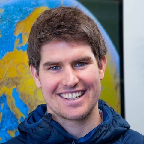Image of Dr Mx Holloway smiling to camera wearing blue hoody against world globe image backdrop