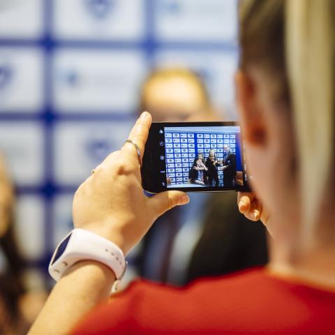 A woman holds a cellphone filming 2 people. BA (Hons) Digital Marketing.