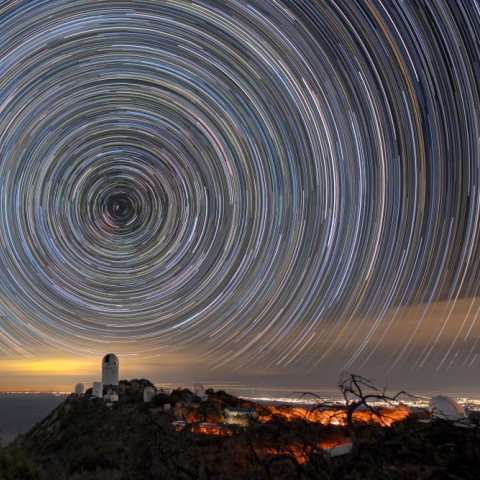 Circles of light on the night sky. A telescope dome atop a mountain is below the center of the circle.
