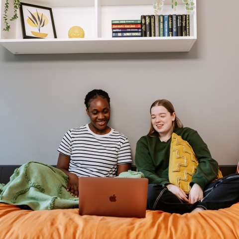 Two students sitting on a bed looking at a laptop