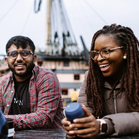 A group of students sitting outside with a ship behind them.