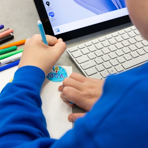 A school pupil working on a drawing with coloured pens, with a laptop in the background