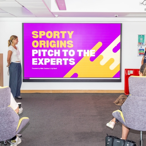 Two female students standing alongside a projector screen as they pitch an idea to industry experts