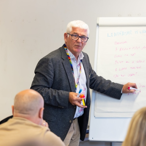 Male with white hair with his hand pointing to a flip chart in a lecture room