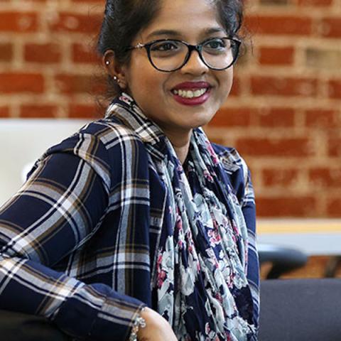Female student in a checkered shirt smiling, sat at a table in front of a brick wall