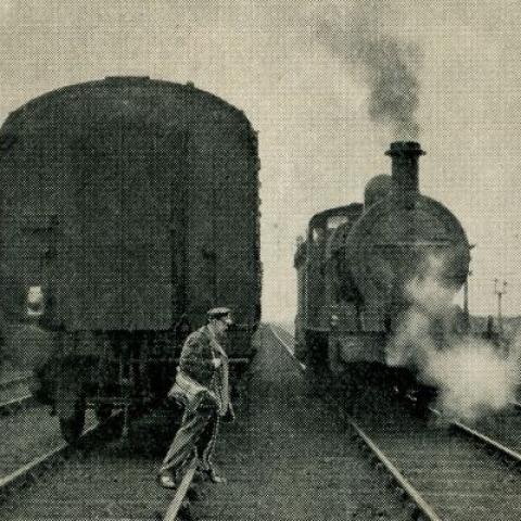 Black and white photo of man crossing railway tracks in front of two trains.
