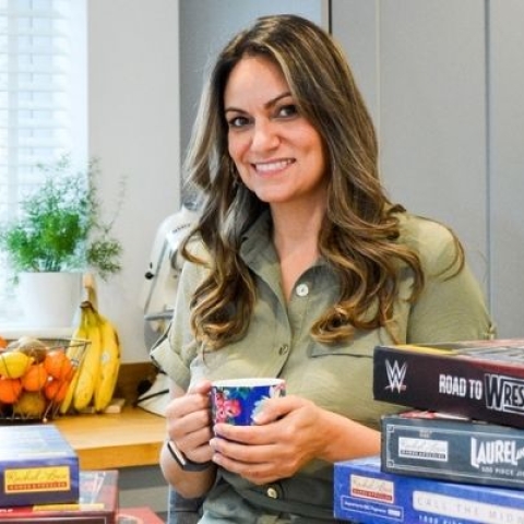 Alumnus Rachel Lowe stood in kitchen with a cup of tea surrounded by the board games she has designed