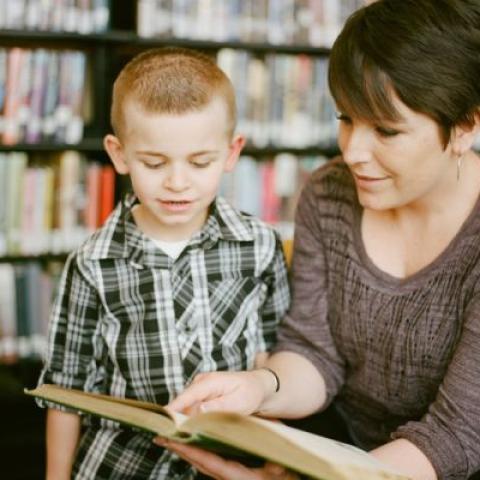 a female teacher with short dark hair and a young boy reading from a hardback book