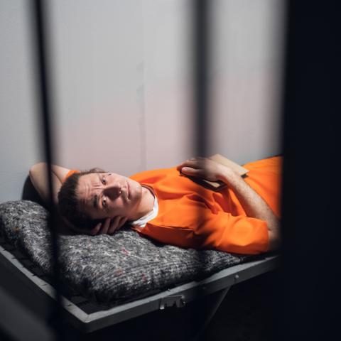 A prisoner in an orange jumpsuit resting on a bunk in a cell with a book. BSc Hons Criminology with Psychology.