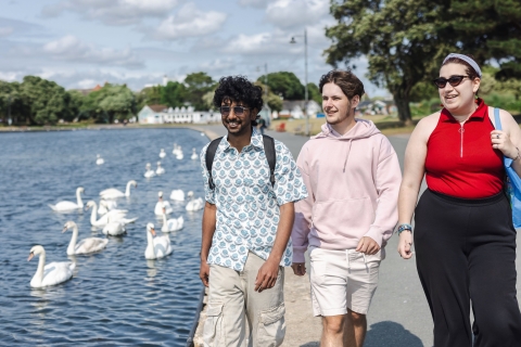 Three happy students walking by a duck pond in Portsmouth, there are swans on the pond, the sun is shining.