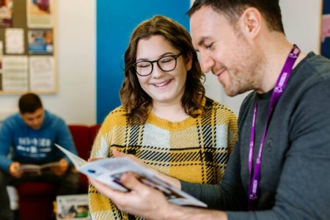 A student with brown hair, glasses and a yellow jumper is listening to student finance advisor James Quinn (who has short brown hair, a grey jumper and a purple lanyard) who is showing her a leaflet