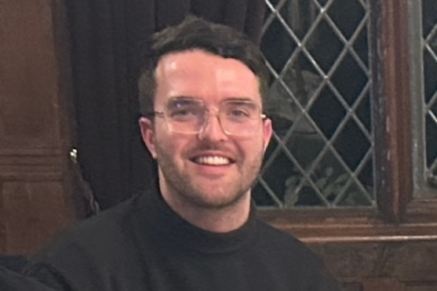 Liam smiling at the camera, wearing a dark jumper, with a window in the background.