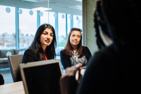 Two females seated in an office talking to person with back to camera.