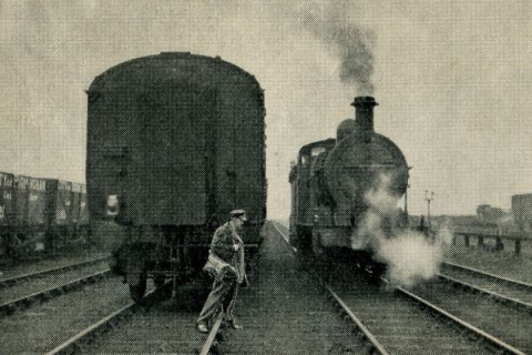 Black and white photo of man crossing railway tracks in front of two trains.