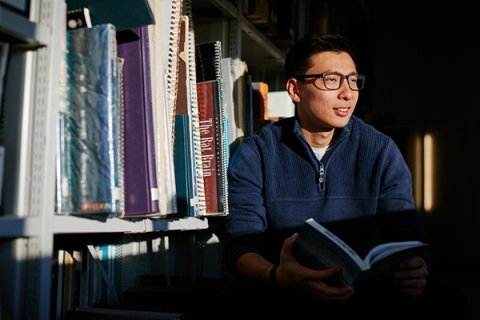 Student holding book next to bookshelf in library