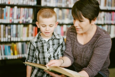 a female teacher with short dark hair and a young boy reading from a hardback book