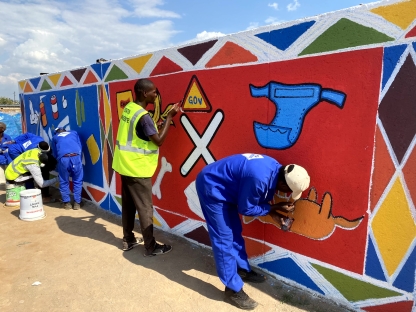 A group of people in South Africa painting a nearly finished mural of recyclable and non recyclable items on a wall. The mural uses lots of bright colour and is extremely eye catching
