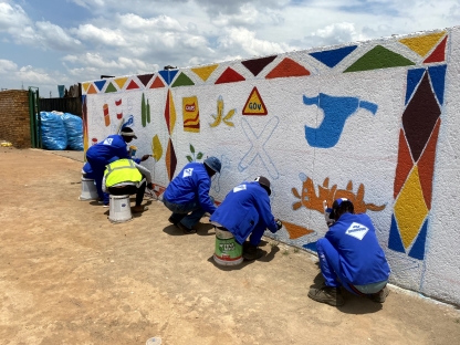 A group of people in South Africa painting a mural of recyclable and non recyclable items. The wall has all items drawn out and the group are now filling the drawings in using bright and eye catching colours 