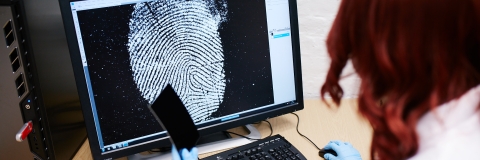 A student with red hair wearing a lab coat, examining fingerprints on a computer.