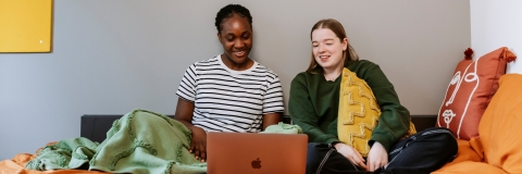 Two students sitting on a bed looking at a laptop