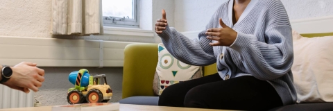 A young woman sat on a chair speaking to someone, next to a a table with a toy on it.