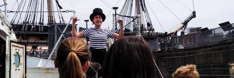 A female in black top hat and black and white striped T-shirt, standing atop a ship deck and flexing her arms to a small crowd of children in school uniform