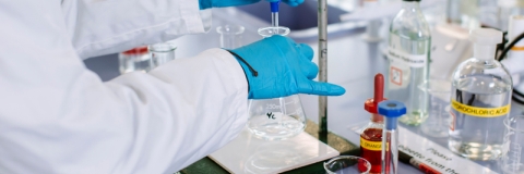 Person in white coat and blue gloves putting liquid in a glass beaker in a lab