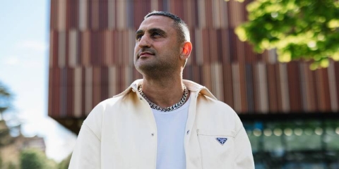 Moses Rashid standing in front of Ravelin Sports Centre looking off into distance in cream prada jacket, white t-shirt and chain necklace