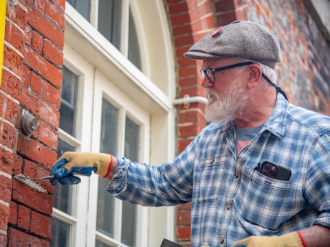 An older man standing to the right of the image in a blue shirt and grey flat cap is putting mortar into a section of old red brick wall to the left. Photo by James Wiley.
