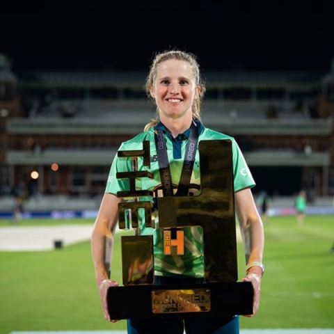 Emily Windsor holding Hundred Cricket trophy on pitch smiling to camera in green kit