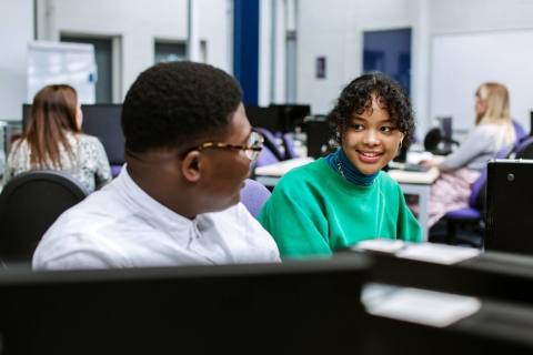 Male and female student sat next to each other talking while working on computer screens