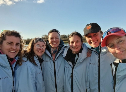 A team of six female rowers smiling on dry land after a training weekend