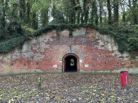 The entrance to a tunnel at Hilsea Lines, there is a large brick wall surrounding the entrance and some overgrown ivy and trees above it. You can just see the light at the other end of the tunnel