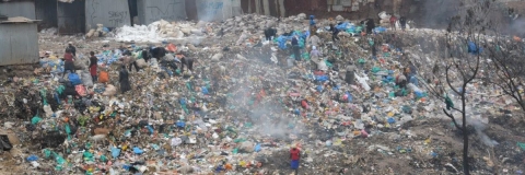 Huge piles of discarded plastic in the Global South - people who look small compared to the piles are walking through it 