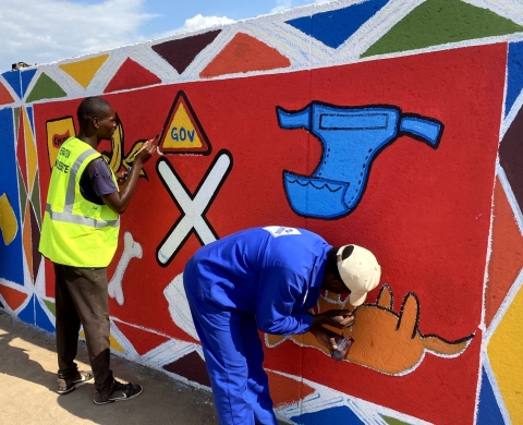A group of people in South Africa painting a nearly finished mural of recyclable and non recyclable items on a wall. The mural uses lots of bright colour and is extremely eye catching