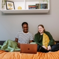 Two students sitting on a bed looking at a laptop