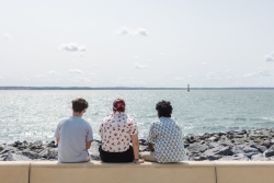 Three students sat on a wall by the sea, backs to the camera