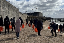 Group walking along a pebble beach carrying orange bags during a coastal clean-up.