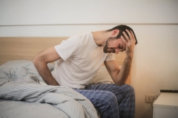 Man sitting on bed with one hand on his head and one on his head, looking in pain