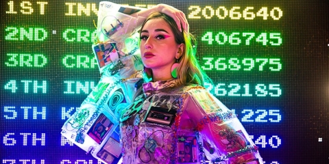 A female person posing by a colourful LCD scoreboard, wearing a plastic jacket decorated with floppy disks and C-90 cassettes