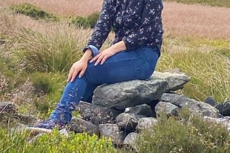 Female wearing navy jeans and navy shirt sitting on a rock with green hills behind her
