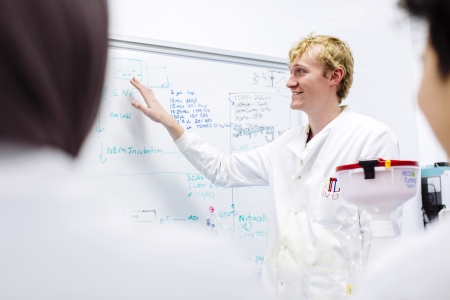 Robert in lab coat explaining something and gesturing to whiteboard