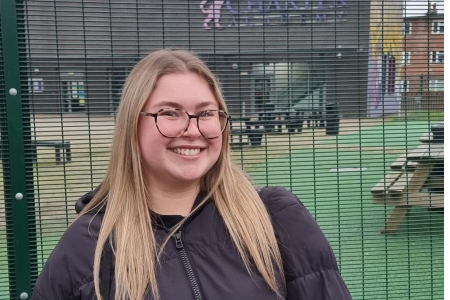 Thea Carpenter - student with long blonde hair, dark black glasses and a black jacket smiling to camera