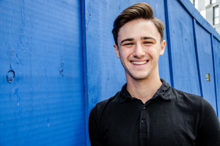 Josh Robinson smiling to camera wearing black t-shirt against blue wall