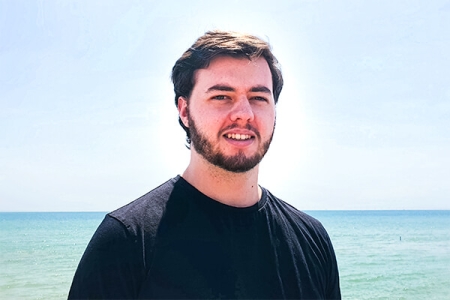 Luke wearing a black T-shirt, on a sunlit beach, standing in front of the sea