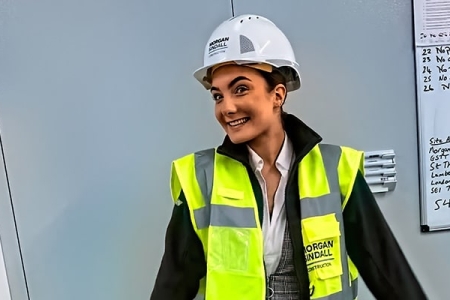 A woman in viz jacket and hard hat, standing in front of a whiteboard with writing on it, smiling at the camera