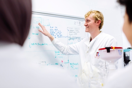 Robert in lab coat explaining something and gesturing to whiteboard